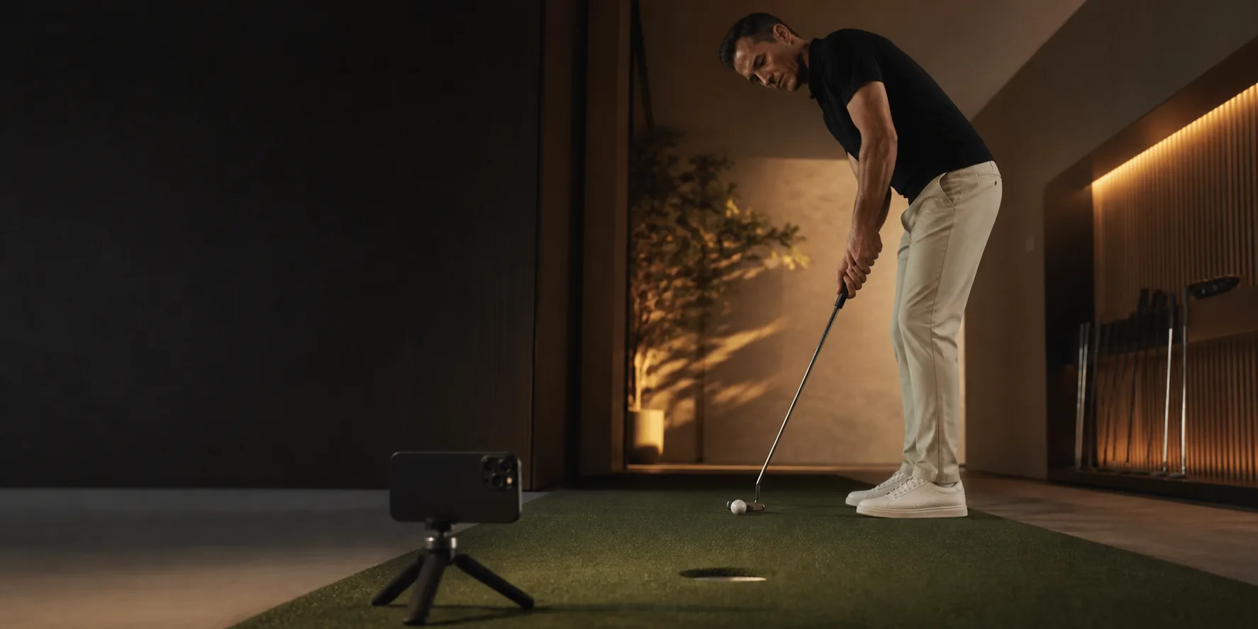 A golfer practicing putting indoors with a phone on a small tripod